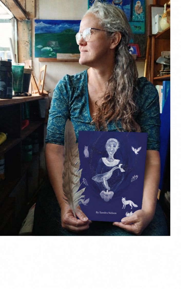 Woman wearing glasses holds a blue artwork featuring a seated figure, cats, and butterflies in a studio.